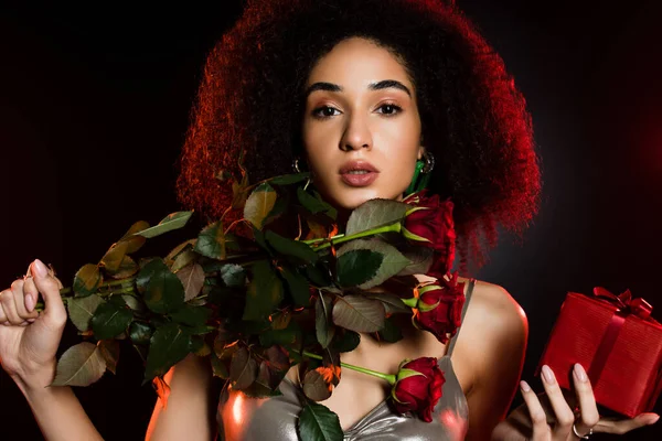 Mujer afroamericana rizada sosteniendo caja de regalo y rosas aisladas en negro - foto de stock