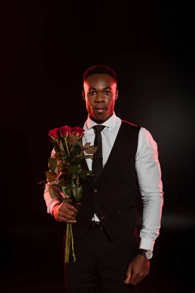 Stylish african american man in formal wear with vest holding red roses on black — Stock Photo