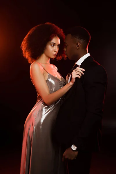 Curly african american woman in dress talking off blazer on boyfriend in suit on black — Stock Photo