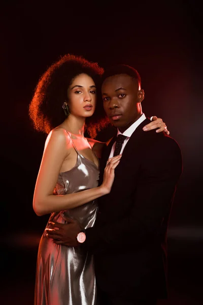 Rizado africano americano mujer en vestido abrazando novio en traje en negro - foto de stock