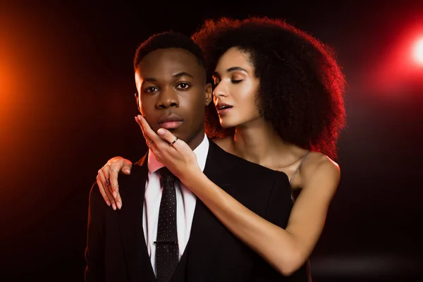 Frisé afro-américain femme toucher visage de copain en costume sur noir — Photo de stock