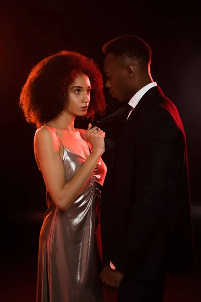 Curly african american woman in dress pulling tie of boyfriend in suit on black — Stock Photo