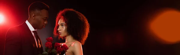 Encaracolado afro-americano mulher com rosas vermelhas perto namorado de terno em preto, banner — Fotografia de Stock