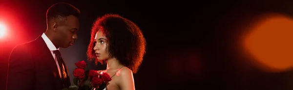 Mujer afroamericana rizada con rosas rojas cerca de novio en traje en negro, pancarta - foto de stock