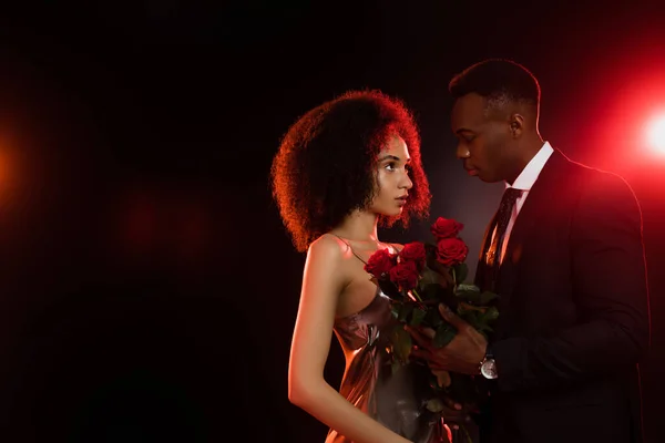 Encaracolado afro-americano mulher com rosas vermelhas olhando para namorado de terno em preto — Fotografia de Stock