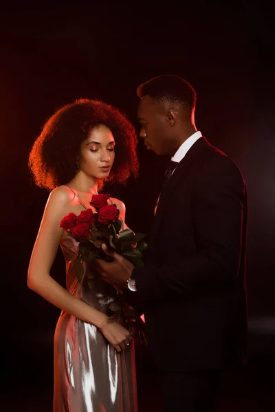 African american man presenting red roses to girlfriend in dress on black — Stock Photo