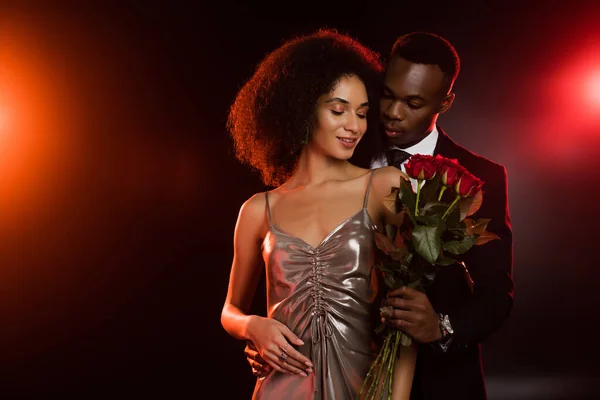 African american man giving red roses to curly girlfriend in dress on black — Stock Photo
