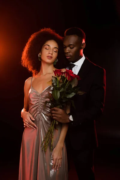 African american man giving red roses to girlfriend in dress on black — Stock Photo