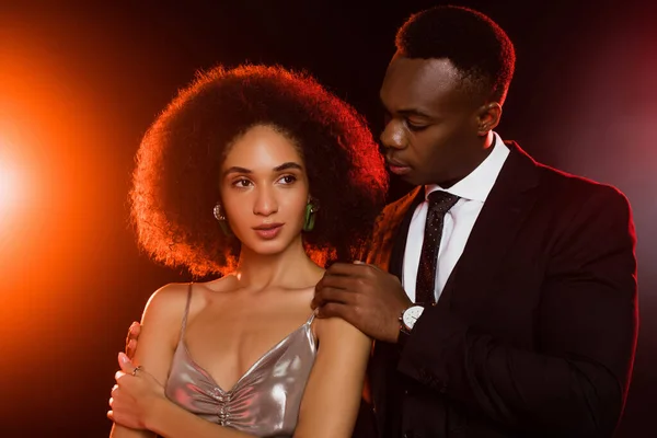 Afro-americano homem no terno abraçando encaracolado mulher no vestido no preto — Fotografia de Stock