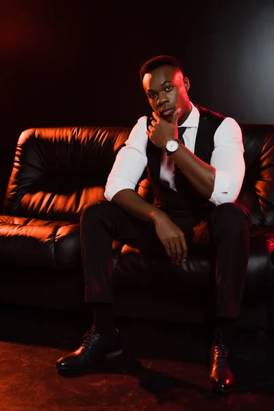 Full length of african american man in suit with vest and tie sitting on sofa on black background — Stock Photo