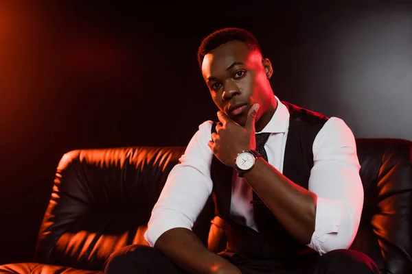 Serious african american man in suit with vest and tie sitting on sofa on black background — Stock Photo