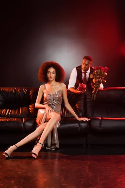 African american man holding red roses and gift box near elegant woman sitting on sofa and black background — Stock Photo