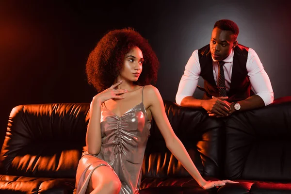 African american man looking at elegant woman sitting on sofa and black background — Stock Photo