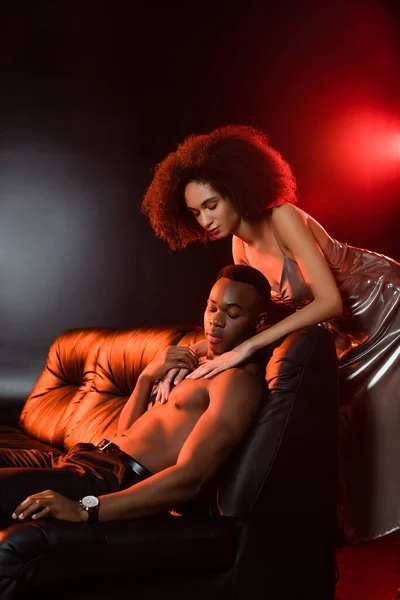 Sexy african american woman in dress seducing shirtless man on sofa and black background — Stock Photo