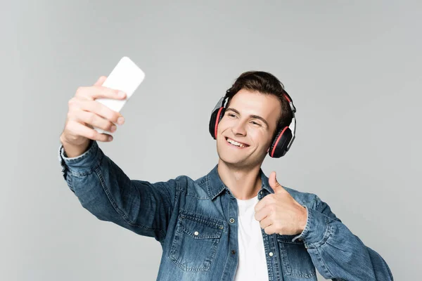 Homem alegre em fones de ouvido mostrando como ao tomar selfie com smartphone isolado em cinza — Fotografia de Stock