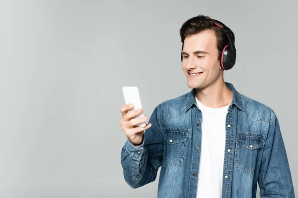 Homme joyeux en jean veste en utilisant smartphone et écouteurs isolés sur gris — Photo de stock