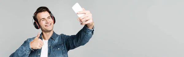 Lächelnder Mann zeigt sich wie beim Selfie mit Smartphone und Kopfhörer isoliert auf grauem Banner — Stockfoto