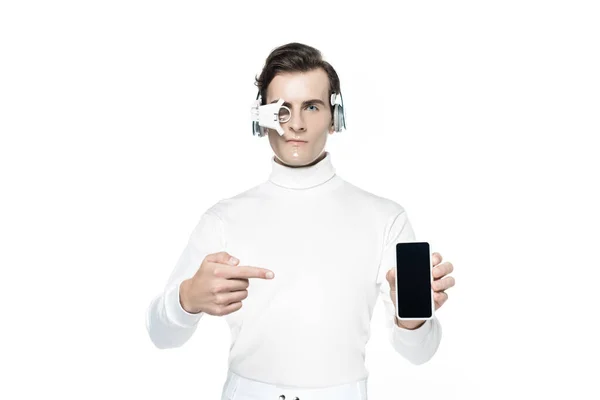 Киборг в глазных линзах и наушниках, указывающих пальцем на смартфон с чистым экраном, изолированным на белом — стоковое фото