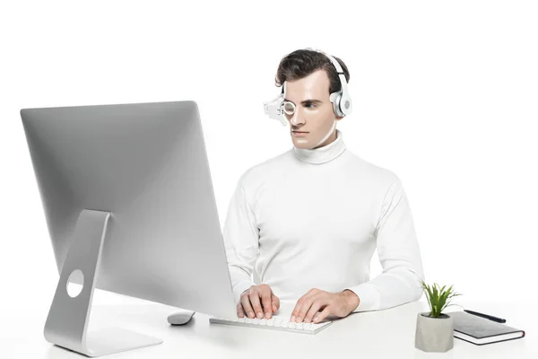 Cyborg in eye lens and headphones using computer near plant and notebook isolated on white — Stock Photo