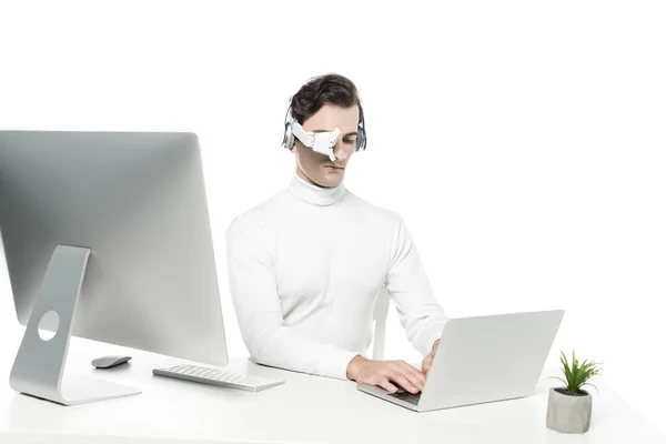 Cyborg en lente de ojo y auriculares usando portátil cerca de la computadora y la planta en primer plano borroso aislado en blanco - foto de stock