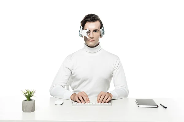 Cyborg in headphones and eye lens using computer keyboard near plant and notebook isolated on white — Stock Photo