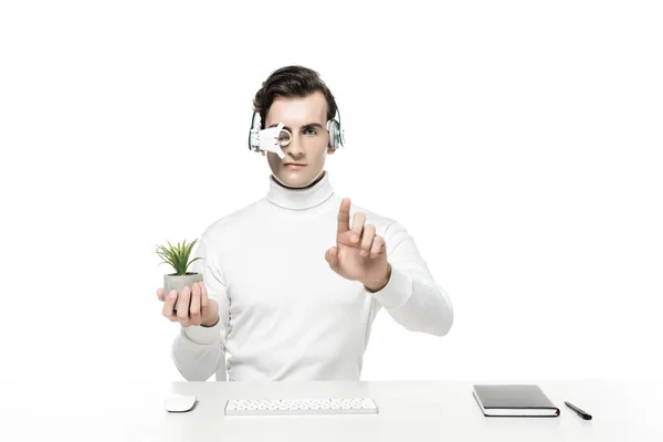 Cyborg en auriculares que sostienen la planta y tocar algo cerca del teclado de la computadora, ratón y portátil aislado en blanco - foto de stock