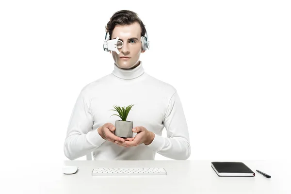 Cyborg em fones de ouvido e lente olho segurando planta perto do mouse computador, teclado e notebook isolado em branco — Fotografia de Stock