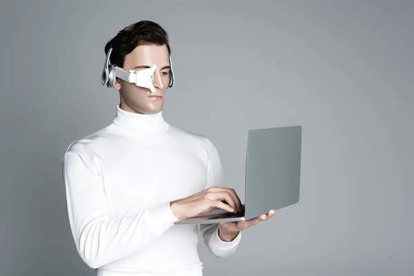 Cyborg in eye lens and headphones using laptop isolated on grey — Stock Photo