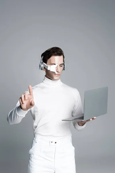 Cyborg hombre en los auriculares con el ordenador portátil y tocar algo aislado en gris - foto de stock