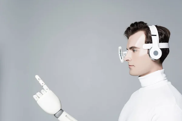 Brunette cyborg in eye lens, headphones and artificial hand pointing with finger isolated on grey — Stock Photo