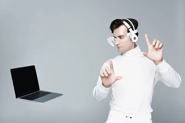 Cyborg dans les écouteurs toucher quelque chose et regarder un ordinateur portable avec écran blanc dans l'air isolé sur gris — Photo de stock