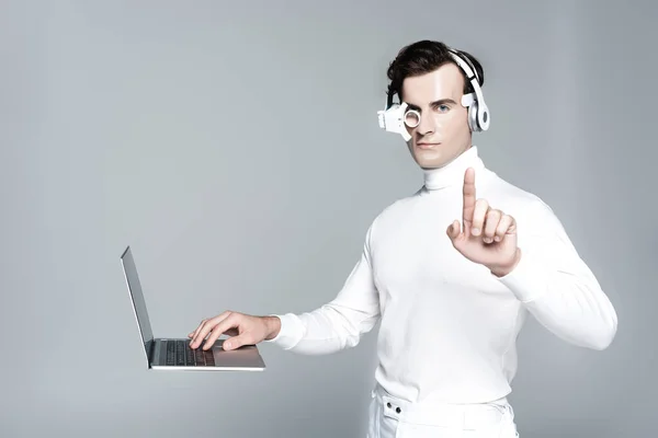 Cyborg in headphones touching something near laptop in air isolated on grey — Stock Photo