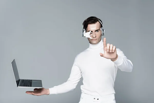 Cyborg mit Kopfhörern, die etwas in der Nähe von Laptop berühren, mit leerem Bildschirm, der in der Luft schwebt, isoliert auf grau — Stockfoto