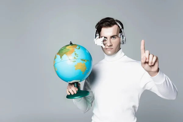 Cyborg in headphones holding globe while pointing up with finger isolated on grey — Stock Photo