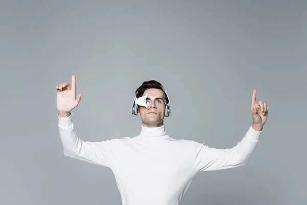 Brunette cyborg man in digital eye lens and headphones looking away while using something isolated on grey — Stock Photo