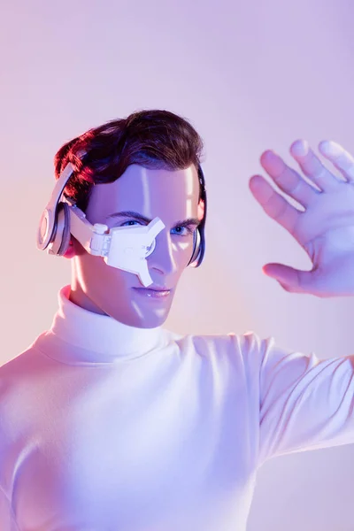 Cyborg man in eye lens and headphones looking at hand on blurred foreground on purple background — Stock Photo