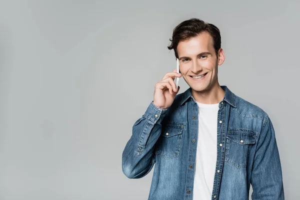 Positive man smiling at camera while talking on smartphone isolated on grey — Stock Photo