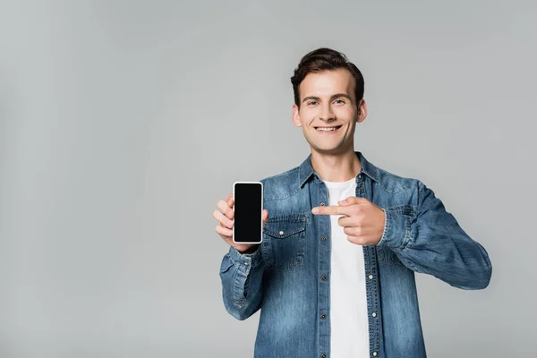 Hombre sonriente apuntando con el dedo al teléfono inteligente con pantalla en blanco aislado en gris - foto de stock