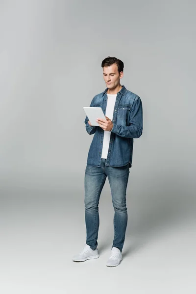 Jeune homme en jean et veste en denim utilisant une tablette numérique sur fond gris — Photo de stock