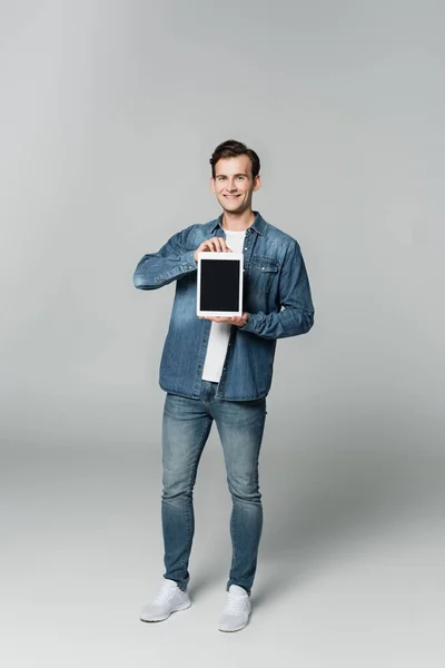 Усміхнений чоловік в джинсовій куртці тримає цифровий планшет з порожнім екраном на сірому фоні — стокове фото
