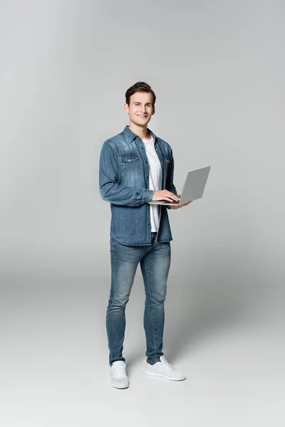 Fröhlicher Mann blickt in die Kamera, während er Laptop auf grauem Hintergrund hält — Stockfoto