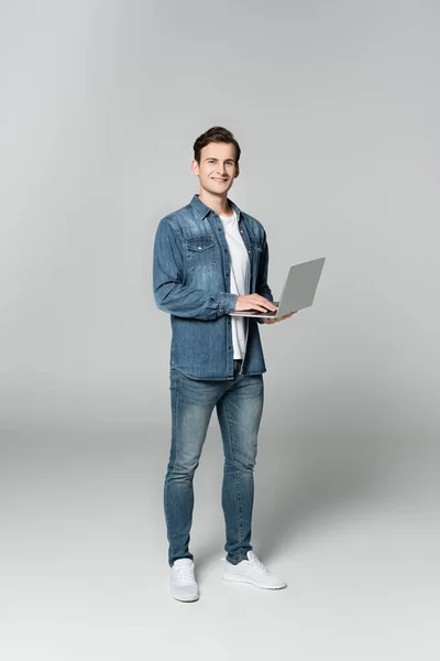 Cheerful man looking at camera while holding laptop on grey background — Stock Photo