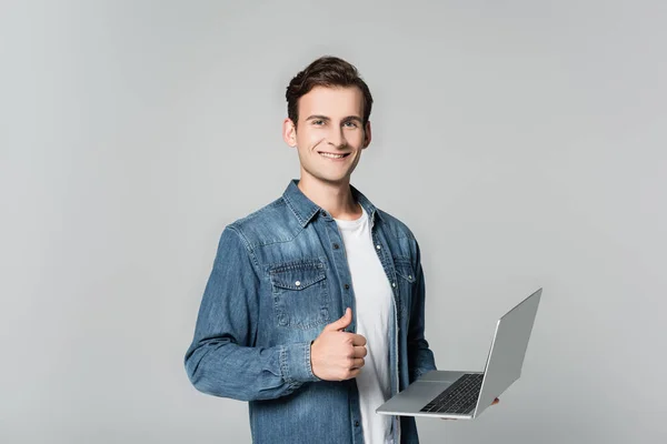 Sonriente hombre mostrando el pulgar hacia arriba mientras sostiene el ordenador portátil aislado en gris - foto de stock