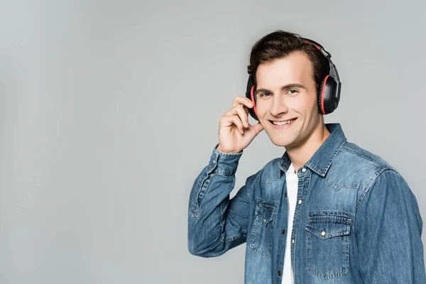 Uomo sorridente in giacca di denim e cuffie guardando la fotocamera isolata su grigio — Foto stock