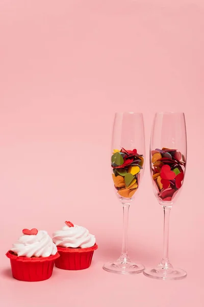 Colorful hearts in champagne glasses and cupcakes on pink — Stock Photo