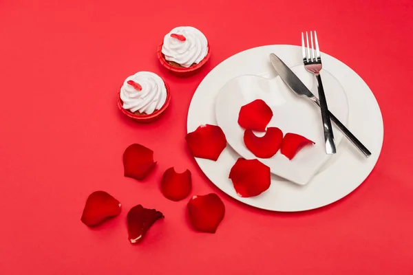 Cupcakes near plate with rose petals and cutlery on red background — Stock Photo