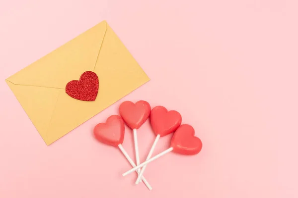 Vista dall'alto di lecca-lecca a forma di cuore e busta con cuore su sfondo rosa — Foto stock