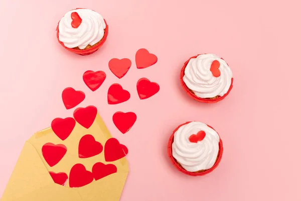 Draufsicht auf rote Herzen, Umschlag und Cupcakes auf rosa — Stockfoto