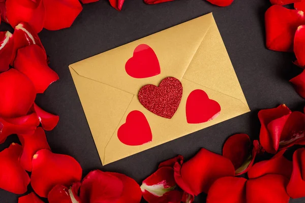 Top view of rose petals and envelope with hearts isolated on black — Stock Photo