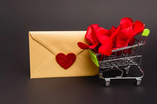 Carrito de compras con pétalos de rosa y sobre aislado en negro - foto de stock